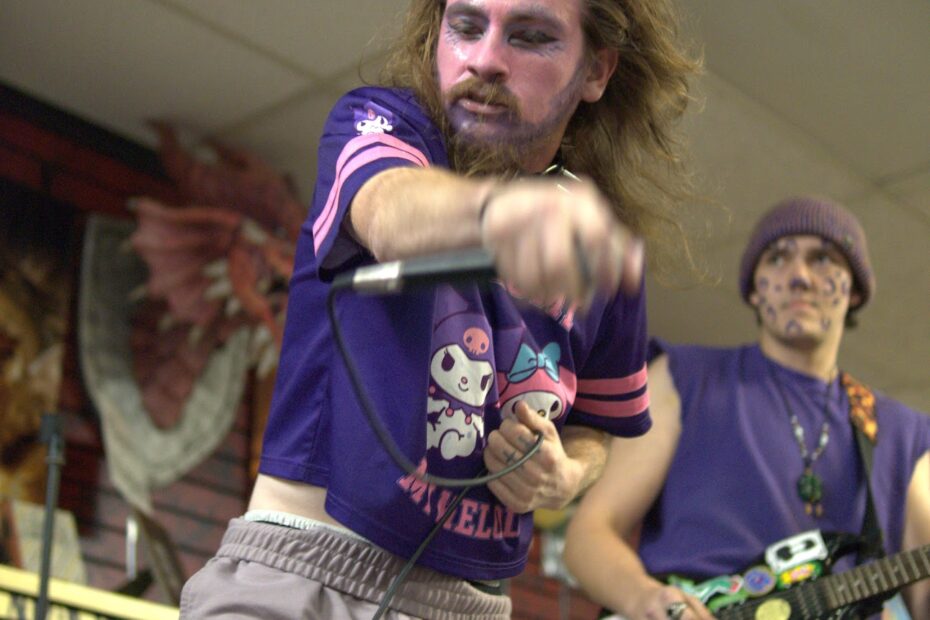A male singer with face paint and a purple My Melody cropped shirt with the words “Melody” visible, singing into a microphone and gesturing with his hand. A second man in the background is playing a black guitar.