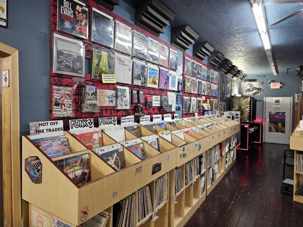 Interior of a record store featuring a long wooden display case of vinyl records, labeled "PUNK", "HARD ROCK & HEAVY METAL", and "ELECTRONIC." Vinyl album covers are displayed on the wall above, with one labeled "BILL EVANS TREASURES," and another reading "HOT OFF THE TRADES" above a record labeled "Marble Madness." The ceiling is blue with black soundproofing tiles, and a bright overhead light runs down the length of the store towards the "EXIT" sign. The floor is dark wood.
